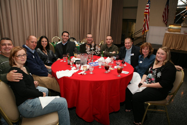 Eine Gruppe von lachenden Menschen sitzt an einem Tisch mit Gläsern, Papieren, Weinflaschen und Flaschen, einer Flagge auf der rechten Seite und Vorhängen an der Wand.