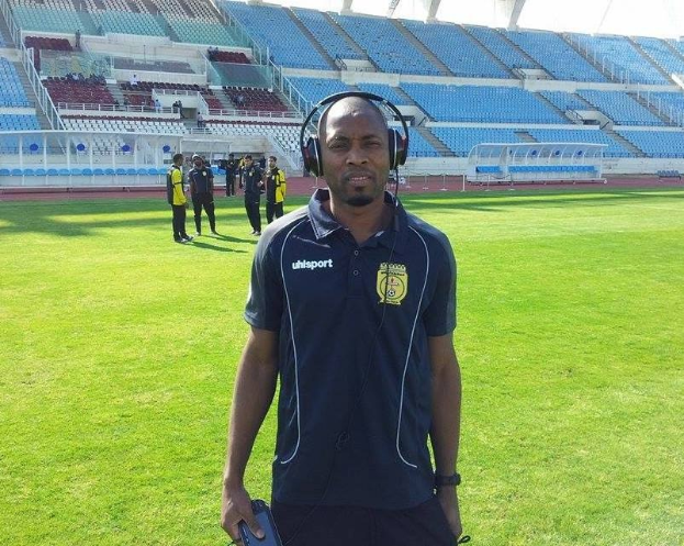 Ein Mann in einem Headset, wahrscheinlich der Trainer der ghanaischen Fußballnationalmannschaft, steht auf einem Fußballfeld in der Nähe eines Stadions und hält ein Objekt in der Hand, während im Hintergrund Menschen zu sehen sind und der Himmel sichtbar ist.