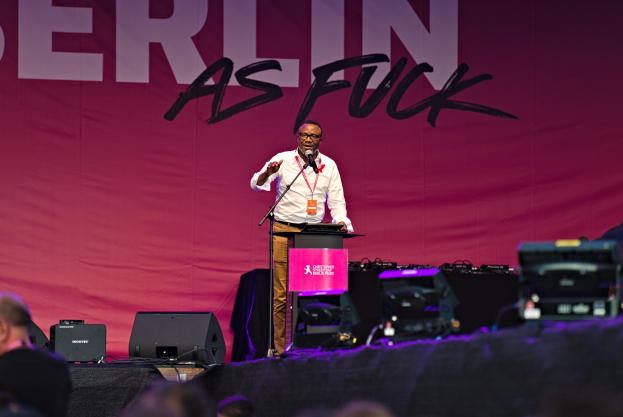 Ein Mann in einem weißen Hemd und braunen Hosen steht auf einer Bühne und hält ein Mikrofon in der Hand, mit einem pinken Banner mit der Aufschrift "Berlin as Fuck" im Hintergrund und sichtbaren Lautsprechern.