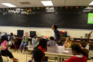 Eine Klasse mit Kindern an Tischen mit verschiedenen Gegenständen, einem Lehrer in der Mitte und einer Tafel im Hintergrund.