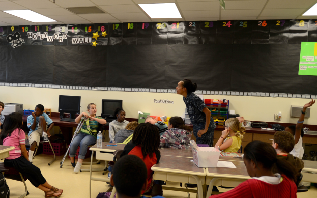 Eine Klasse mit Kindern an Tischen mit verschiedenen Gegenständen, einem Lehrer in der Mitte und einer Tafel im Hintergrund.