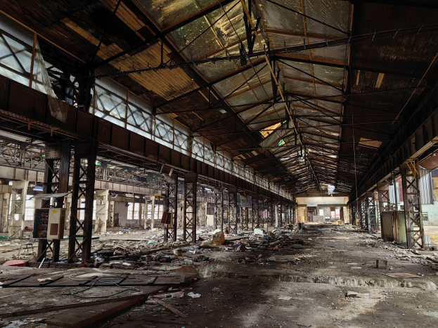 Verlassene Fabrikhalle mit verstreuter Trümmern, kaputten Fenstern, Metallstangen, Deckenlampen und einem beschädigten Dach.