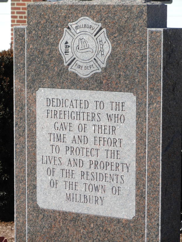 Gedenkstein mit einer Plakette für Feuerwehrleute in Millbury, vor einem Backsteingebäude mit umliegender Vegetation aufgestellt.
