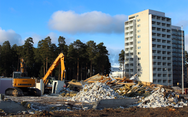 Gro├če Schutthalde vor einem hohen Geb├Ąude auf einer Baustelle, umgeben von Kr├Ąnen, Steinen, Pfosten und B├Ąumen.