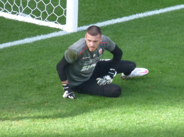 Ein Fußballspieler kniet vor einem Tor auf dem Boden, trägt einen Sportdress, Handschuhe und Turnschuhe und hat den Torpfosten im Hintergrund sichtbar.
