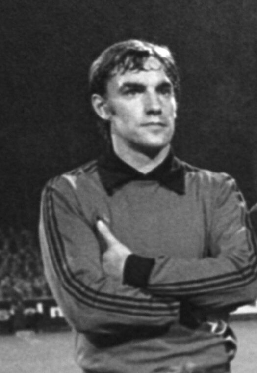 Schwarzes und weißes Foto eines Mannes, der mit verschränkten Armen auf einem Fußballfeld steht, umgeben von einer Gruppe von Menschen im Hintergrund.