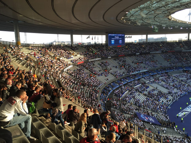 Große Menschenmenge in einem Stadion bei einem Fußballspiel, mit einer Bühne, Fahnen und der Allianz Arena in München, Deutschland im Hintergrund.