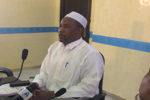 Ein Mann in einem weißen Gewand und einer Kappe sitzt an einem Tisch mit Mikrofonen, Papieren und anderen Gegenständen und spricht, während rechts von ihm eine andere Person mit Brille sitzt, in einem Raum mit einer Wand und einer Tür im Hintergrund.