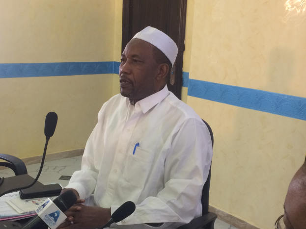 Ein Mann in einem weißen Gewand und einer Kappe sitzt an einem Tisch mit Mikrofonen, Papieren und anderen Gegenständen und spricht, während rechts von ihm eine andere Person mit Brille sitzt, in einem Raum mit einer Wand und einer Tür im Hintergrund.