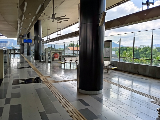 Leerer Flughafen-Terminal mit großem Stadtblick-Fenster, Innenpfeiler, Informationsanzeigen, Deckenleuchten, Deckenventilatoren und Außenblick auf Bäume, Gebäude, Hügel und bewölkten Himmel.