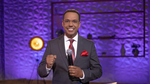 Stephen Colbert, der einen Anzug, Krawatte und Brille tr├Ągt, steht auf einer B├╝hne und h├Ąlt ein Mikrofon in der Hand, mit einer Wand und verschiedenen Gegenst├Ąnden dahinter.