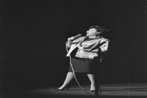 Eine Frau in einem schwarzen und weißen Outfit mit 1950er-Jahre-Frisur singt enthusiastisch in ein Mikrofon auf der Bühne, beleuchtet von einem Scheinwerfer.