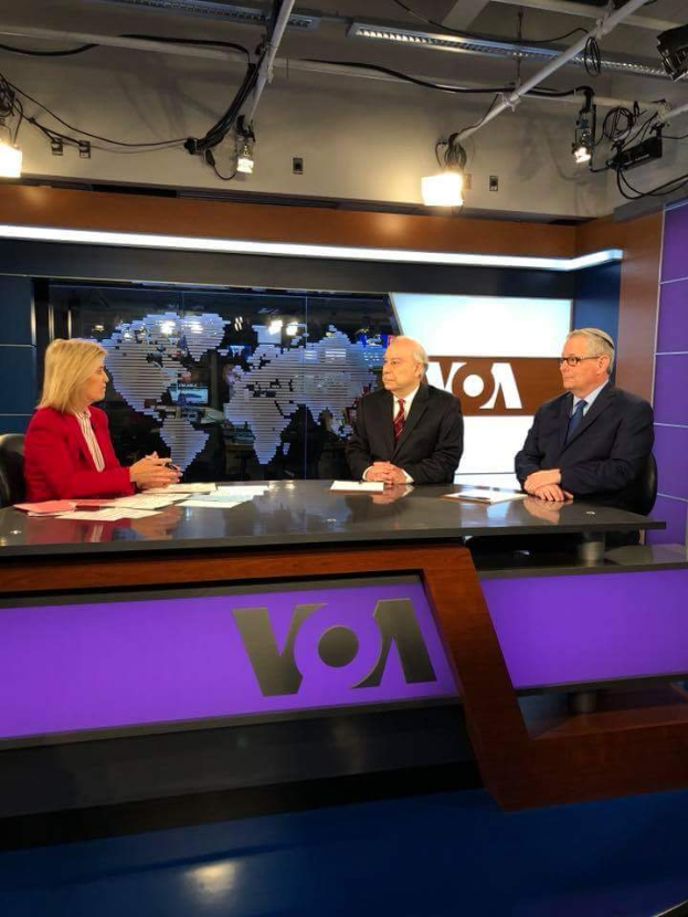 Drei Personen, zwei Männer in Blazern und Krawatten und eine Frau in einem roten Kleid sitzen an einem Schreibtisch mit Papieren und Gegenständen, die in eine Diskussion vor einem Fernseher vertieft sind, mit einem Bildschirm und Lichtern im Hintergrund.