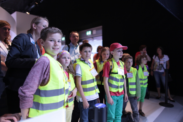 Eine Gruppe von Kindern in Sicherheitswesten und Namensschildern, mit einem Jungen, der links lächelt, auf einem Boden mit einem Pfahl auf der rechten Seite und einigen Menschen im Hintergrund.