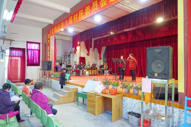 Eine Bühne mit zwei singenden Personen, eine Band im Hintergrund, rote Vorhänge, eine Lampe, ein Lautsprecher rechts und ein Publikum auf Stühlen sitzend.