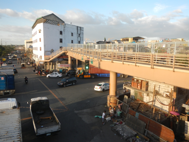 Eine belebte Stadtstraße mit verschiedenen Fahrzeugen, Fußgängern, einer Brücke, Ampeln, Gebäuden, Bäumen und einem bewölkten Himmel.