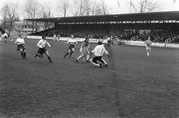 Eine Gruppe von Männern, die auf einem Rasenfeld Hockey spielen, mit einem Schuppen, Zuschauern auf den Tribünen, Bannern, Fahnen, Bäumen, Gebäuden und einem klaren Himmel, dargestellt in Schwarz-Weiß.