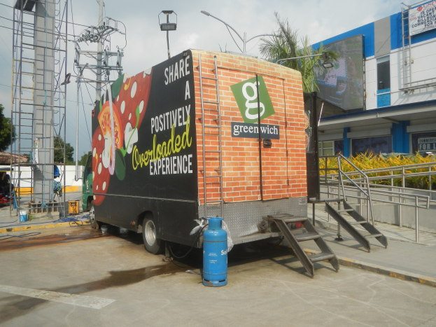 Ein Lkw vor einem Gebäude mit einer Tafel, auf der "Teilen Sie ein positiv überladenes Erlebnis" steht, mit Stufen, einem Zylinder, Geländern, Strommasten mit Kabeln, Laternen, Bäumen und einem bewölkten Himmel.