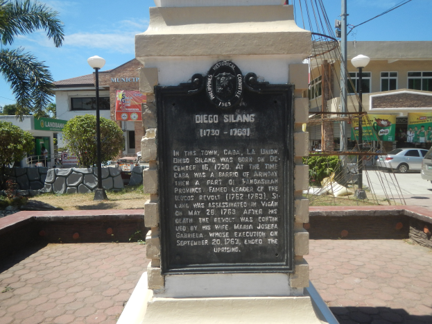 Ein Denkmal mit einer Plakette steht in der Mitte einer Straße, umgeben von Gebäuden, Straßenmasten, Straßenlaternen, Strommasten, Stromkabeln, Kraftfahrzeugen, Büschen, Bäumen und einem bewölkten Himmel.