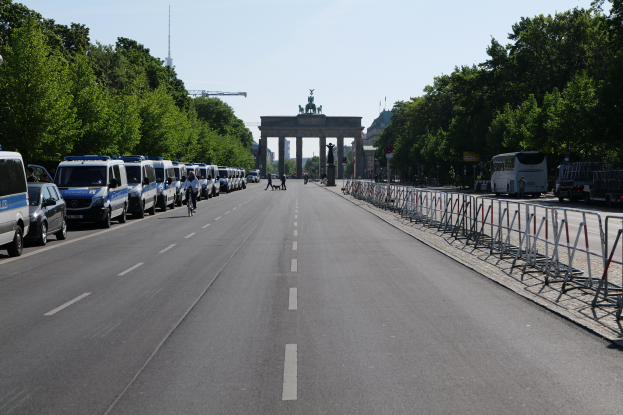 Eine lange Reihe von Polizeiwagen, die auf der Seite einer Straße vor dem Brandenburg-Tor in Berlin, Deutschland, geparkt sind, mit Menschen, die Fahrräder fahren und auf der Straße stehen, sowie Barrieren und Bäume an den Seiten und einem Bogen mit Statuen im Hintergrund.