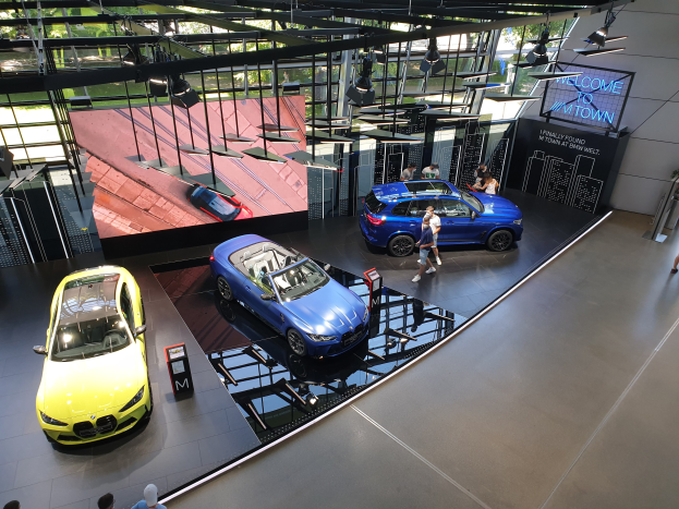 Eine Gruppe von Menschen, die im BMW Museum in München stehen, mit Autos auf der linken Seite und einem Schild mit der Aufschrift "BMW Museum München" auf der rechten Seite, mit Blick auf Büsche, Geböude und den Himmel durch Glasfenster.