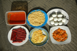 Ein Tisch mit Tellern voller Nudeln, Eiern, Würstchen und einer Schüssel Sauce, ordentlich angerichtet für eine Mahlzeit.