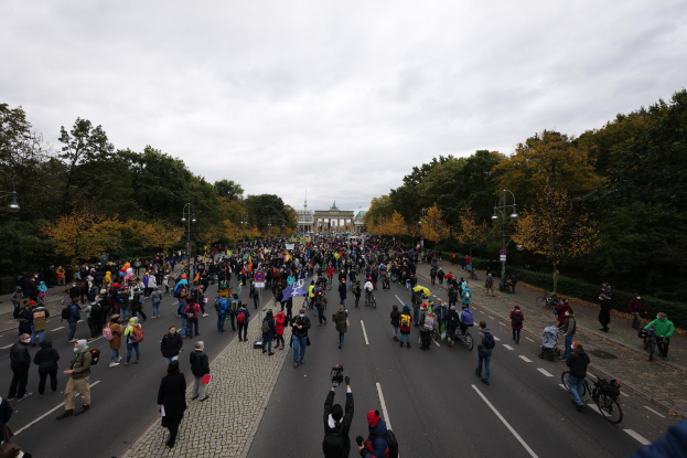 Große Menschenmenge marschiert auf einer baumbestandenen Straße in Berlin, einige halten Kameras, mit einem Gebäude und einem klaren Himmel im Hintergrund.