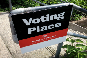Ein Wahlplatzschild an einem Gebäude mit einer Tafel an einem Geländer in der Vordergrund und Pflanzen im Hintergrund.