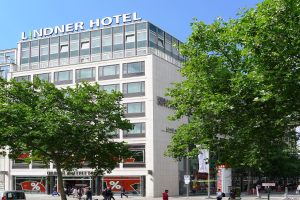 Großes modernes Gebäude mit der Aufschrift "Lindner Hotel" in Berlin, mit Glasfenstern, umgeben von Bäumen und einer belebten Straße mit Fußgängern und Fahrzeugen unter einem sichtbaren Himmel.