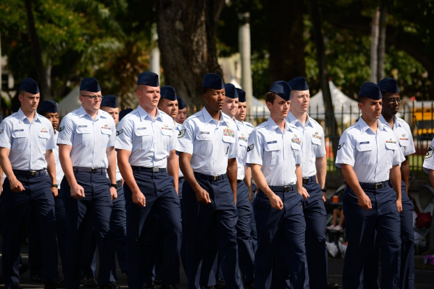Männer in Uniform auf einer Straße mit einem Eisengeländer dahinter, Bäume und Pfosten im Hintergrund.