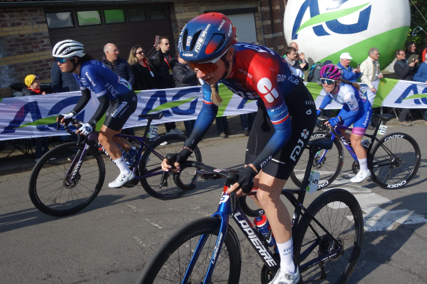 Gruppe von Radfahrern, die eine Straße mit einem Banner im Hintergrund hinunterfahren, einige tragen Helme und halten Kameras, mit einem Gebäude, Bäumen und einem Ballon im Hintergrund.