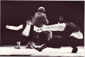 Eine Gruppe von Menschen, die auf einer Bühne tanzen, in einem Schwarz-Weiß-Foto, mit einer Person in der Mitte, die Schuhe trägt, vor einem dunklen Hintergrund.