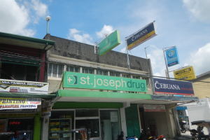 Eine belebte Straßenszene mit verschiedenen Gebäuden, Motorrädern, Geschäften, Schildern und Strommasten, mit einer Apotheke namens "st. joseph drug" im Vordergrund, unter einem bewölkten Himmel.