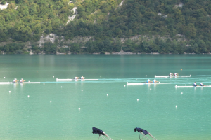 Eine Gruppe von Menschen, die auf einem Gewässer umgeben von Bäumen und einem Hügel rudern, mit Kleiderständen am unteren Bildrand.