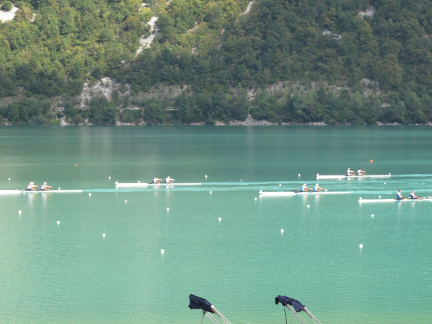 Eine Gruppe von Menschen, die auf einem Gewässer umgeben von Bäumen und einem Hügel rudern, mit Kleiderständen am unteren Bildrand.