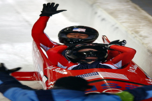 Zwei Bobsleighs fahren eine Bahn herunter, einer mit sichtbarem Helm und Handschuhen, mit einer Person unten und einer Wand im Hintergrund.