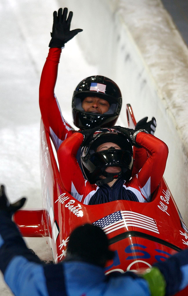 Zwei Bobsleighs fahren eine Bahn herunter, einer mit sichtbarem Helm und Handschuhen, mit einer Person unten und einer Wand im Hintergrund.