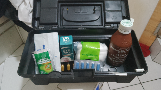 Eine schwarze Box auf dem Boden enth├Ąlt eine Flasche Fl├╝ssigkeit, einige Packungen und andere Gegenst├Ąnde, mit einem wei├čem Tuch links daneben.