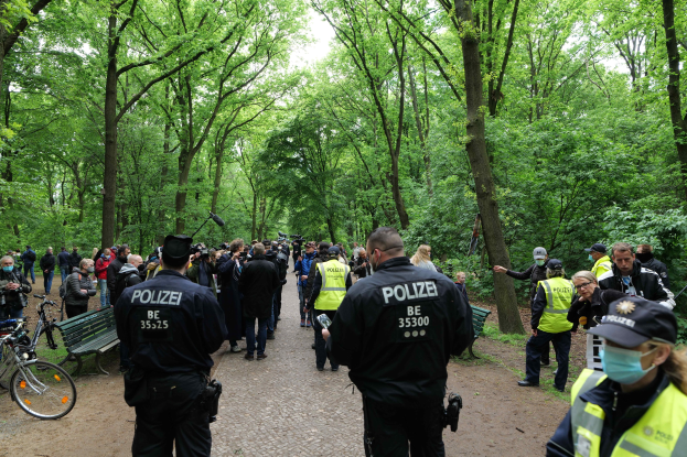 Polizisten vor einer Menschenmenge bei einer Outdoor-Veranstaltung, mit Fahrrädern und einer Bank im Vordergrund und Bäumen im Hintergrund.