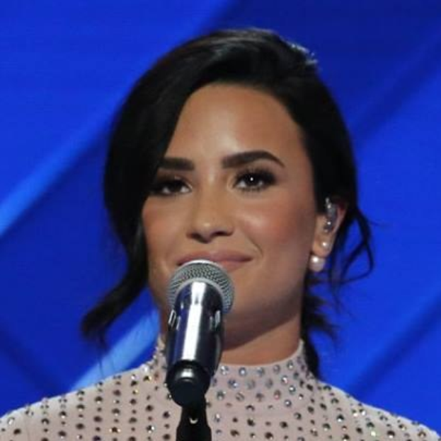 Demi Lovato steht selbstbewusst vor einem Mikrofon auf der Grammy-Bühne, trägt ein schwarzes Kleid und einen eleganten Bob, mit einem blauen Hintergrund.