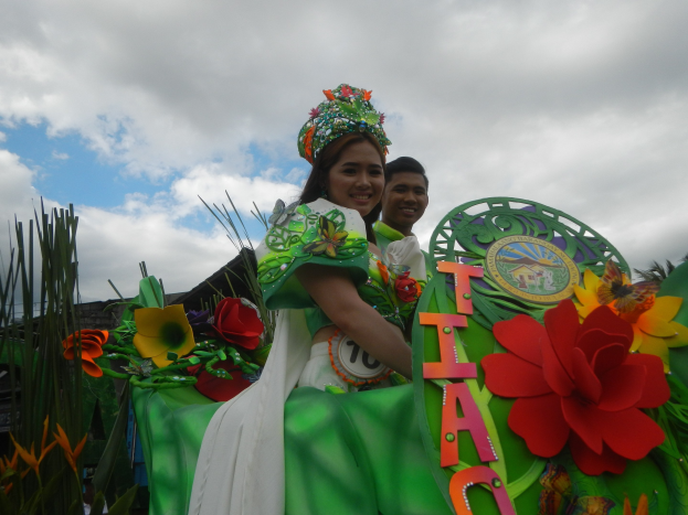Zwei Personen auf einem grün-weißen Paradewagen, die Frau in einem grünen Kleid und der Mann hinter ihr stehend, mit Blumenschmuck und Text auf dem Wagen vor einem Hintergrund aus Bäumen und einem klaren Himmel.