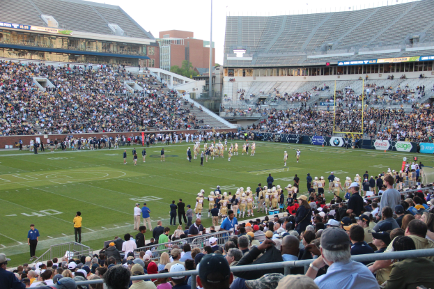 Ein Fußballspiel in einem Stadion mit Spielern auf dem Feld und Zuschauern in den Rängen unter einem klaren blauen Himmel.