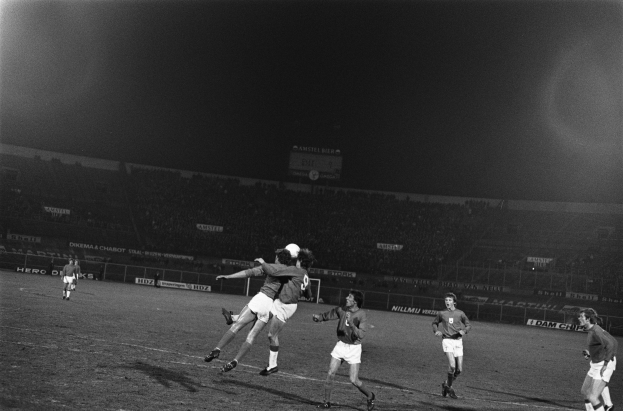 Schwarzes und weißes Bild von Männern, die auf einem Feld Füssball spielen, mit Zuschauern auf den Tribünen und einer Werbetafel im Hintergrund.