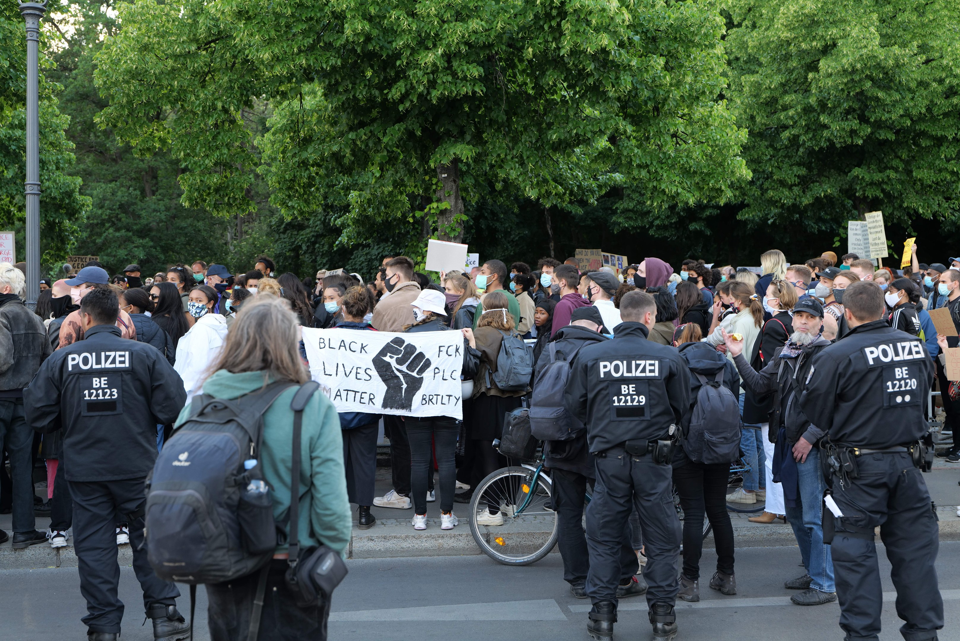 Eine große Gruppe von Menschen nimmt an einer Black-Lives-Matter-Demonstration in Berlin teil, einige halten Schilder und tragen Mützen und Taschen, während im Vordergrund ein Fahrrad und im Hintergrund Bäume und ein Pfahl zu sehen sind.