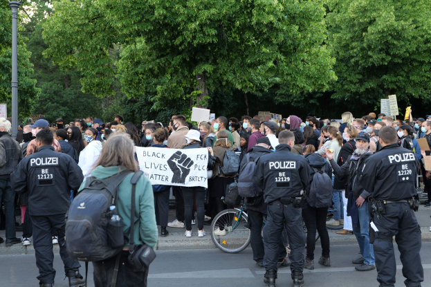 Eine große Gruppe von Menschen nimmt an einer Black-Lives-Matter-Demonstration in Berlin teil, einige halten Schilder und tragen Mützen und Taschen, während im Vordergrund ein Fahrrad und im Hintergrund Bäume und ein Pfahl zu sehen sind.