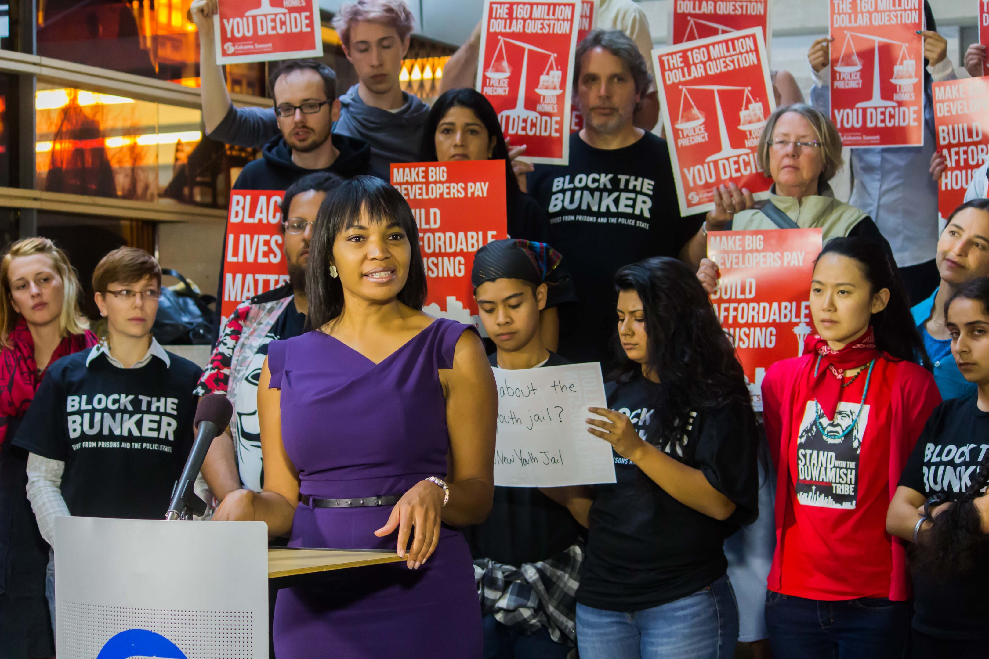 Eine Black-Lives-Matter-Aktivistin spricht in ein Mikrofon auf einem Podium, wo sie eine Menge mit Schildern anspricht, mit Scheinwerfern und Equipment im Hintergrund.