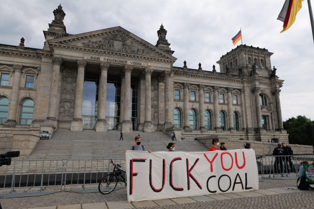 Eine Gruppe von Menschen mit einem Transparent mit der Aufschrift "Fuck You Coal" vor dem Reichstaggebäude in Berlin, Deutschland, mit einem Fahrrad in der Nähe, Bäumen, einem Fahnenmast und einem bewölkten Himmel.