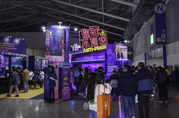 Eine große Gruppe von Menschen steht vor einem Messestand, der als 'Indie Game Development Award 2019' gekennzeichnet ist, wobei einige Taschen und verschiedene Anzeigen, Bildschirme und Lichter im Hintergrund zu sehen sind.