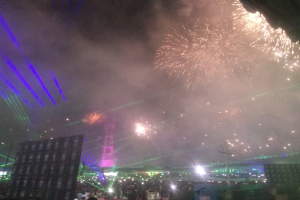 Eine Menschenmenge bei einem Konzert mit beleuchteten Gebäuden und Masten im Hintergrund, unter einem Himmel voller bunter Feuerwerke.