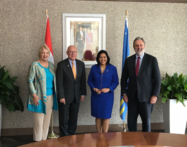 Gruppe von Menschen, die um einen Tisch mit Papieren stehen, umgeben von Fahnen, Topfpflanzen und einem Rahmenfoto an der Wand während einer Unterschriftszeremonie.